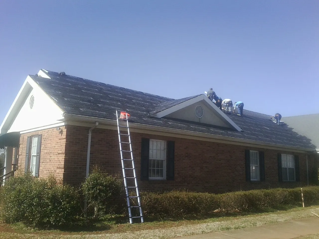 Crew tearing shingles off a brick ranch roof with a ladder set — Metal Roof Repair work by our crew in Stacyville