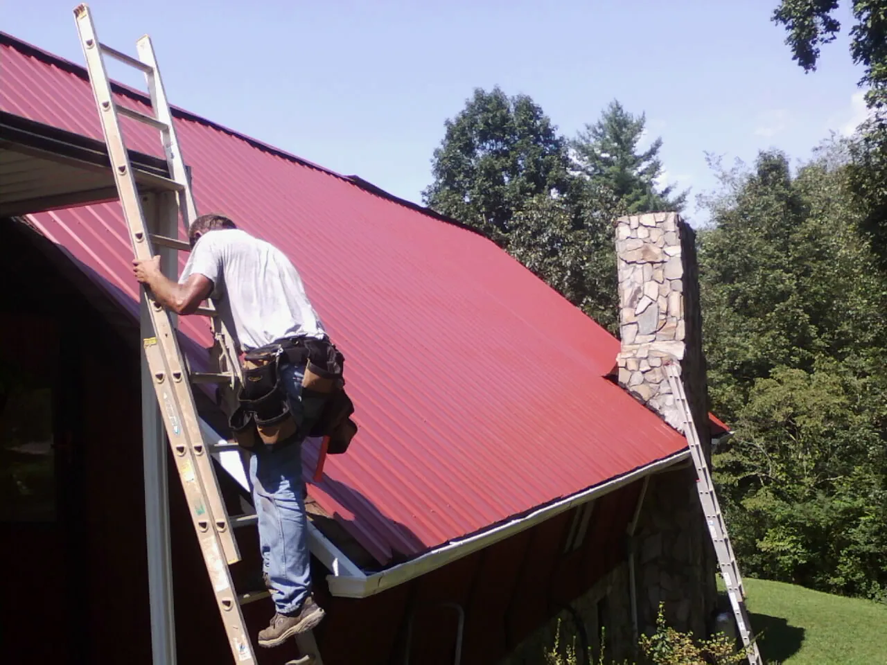 Roofer on a ladder beside a red metal roof and stone chimney — Soffit & Fascia Repair by Roofing Brothers in Stacyville